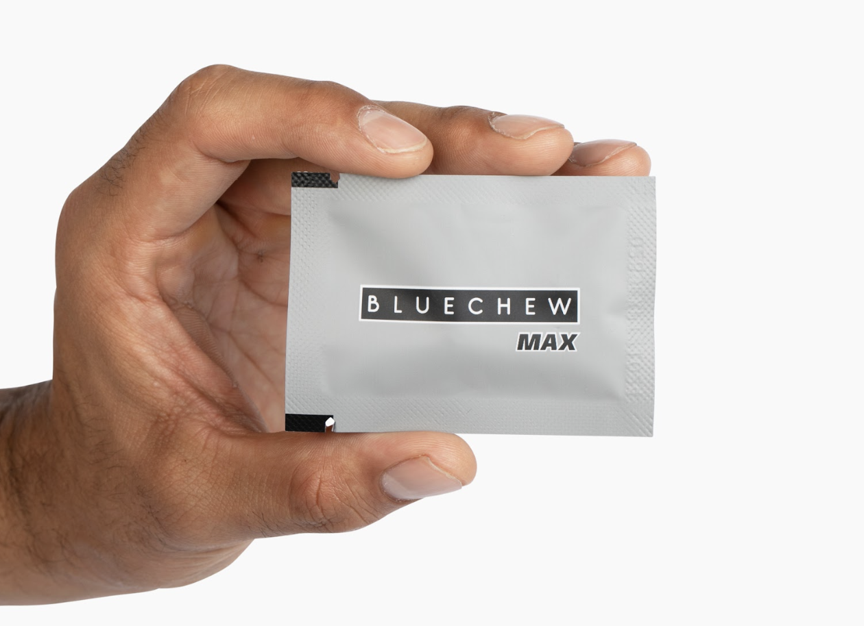 A hand holding a packet of BlueChew MAX, which features a gray design with the brand name prominently displayed.
