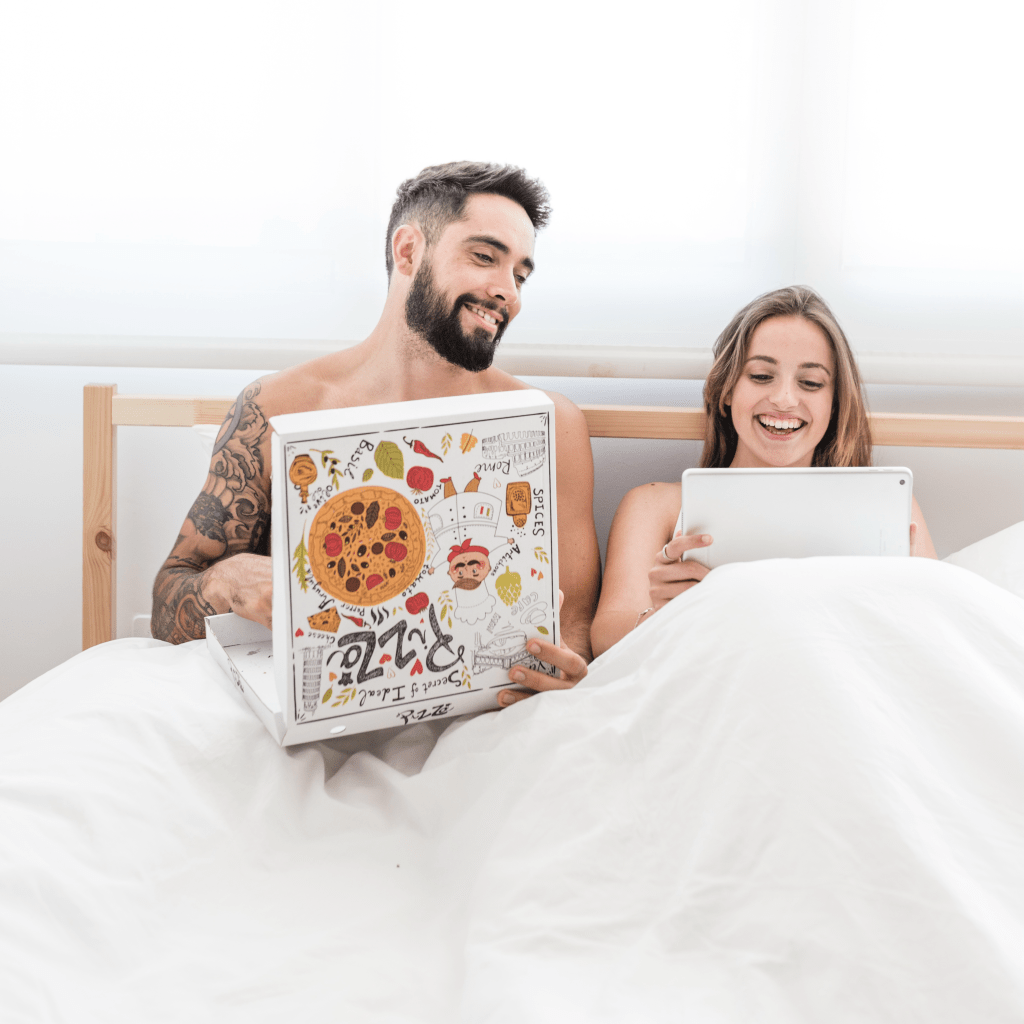 A happy couple laying in bed together, smiling and enjoying each other's company, with one holding a pizza box.