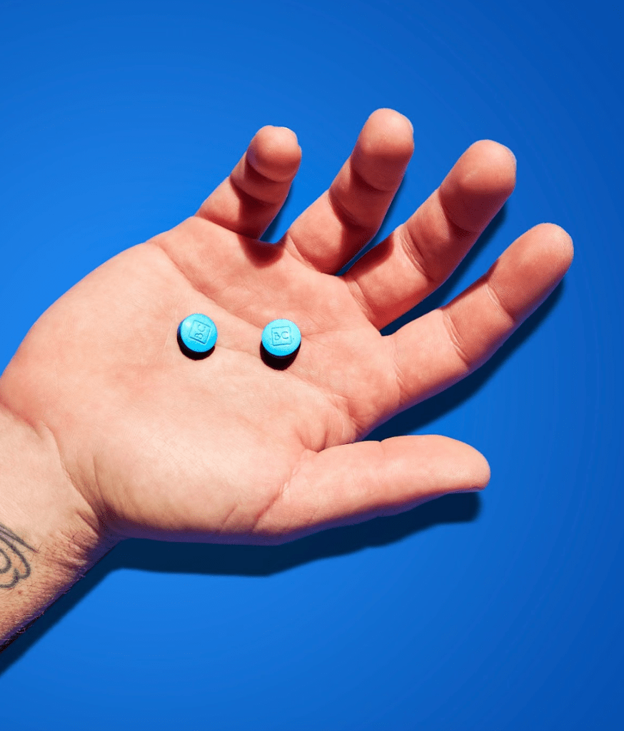A man's hand holding two bluechew tablets against a blue background.