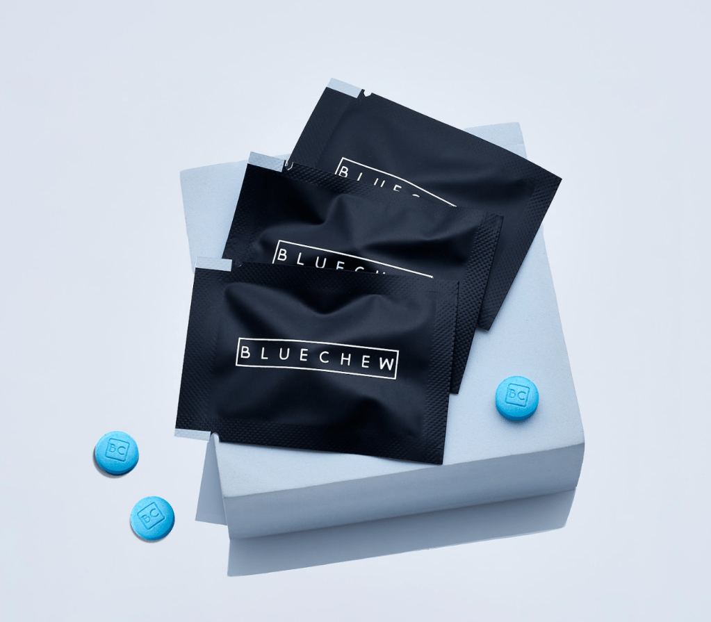 Three packets of BlueChew erectile enhancement medication and two blue tablets displayed on a light-colored surface.