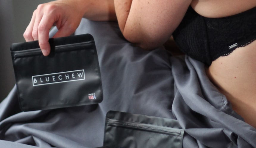 A man holding a Black pouch labeled 'BLUECHEW' while laying in a bed with a woman with grey sheets.